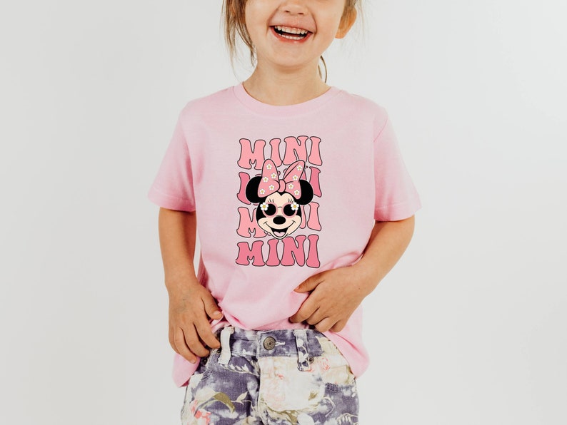 K&ouml;nnte beinhalten: Ein pinkfarbenes T-Shirt mit einem Aufdruck von Minnie Mouse, die eine Sonnenbrille und eine Schleife mit Blumen tr&auml;gt. Der Text "MINI" wird viermal auf dem Shirt wiederholt.