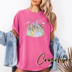 May include: A magenta t-shirt featuring a colorful graphic of Disney princesses in front of a castle with fireworks. The shirt is worn with a light blue denim jacket. The word "Crunchberry" is written in a cursive font.