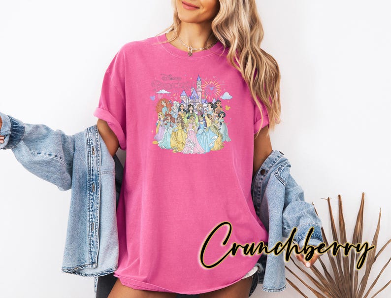 May include: A magenta t-shirt featuring a colorful graphic of Disney princesses in front of a castle with fireworks. The shirt is worn with a light blue denim jacket. The word "Crunchberry" is written in a cursive font.