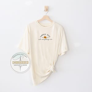 May include: Cream-colored t-shirt with the text "Pumpkin Spice and everything nice" in black script and a small orange pumpkin graphic. The shirt is hanging on a wooden hanger against a white wall.