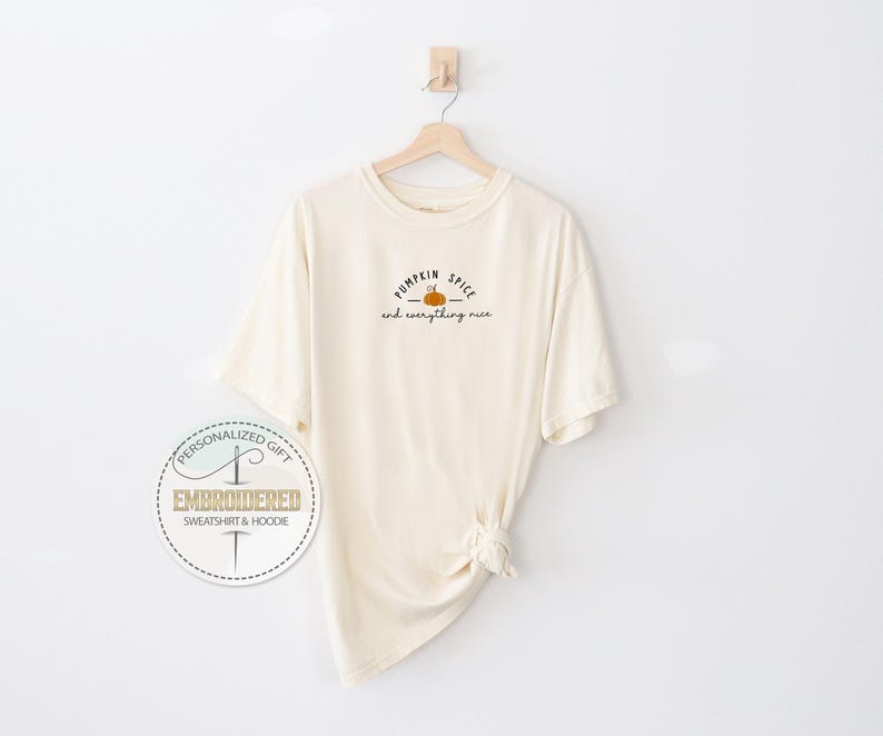 May include: Cream-colored t-shirt with the text "Pumpkin Spice and everything nice" in black script and a small orange pumpkin graphic. The shirt is hanging on a wooden hanger against a white wall.