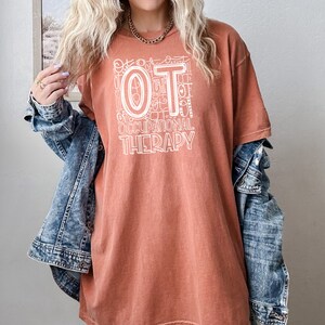 May include: A rust-colored t-shirt with a white graphic design that says "OT Occupational Therapy". The graphic is made up of the letters "OT" surrounded by smaller versions of the letters "OT" and other related words.