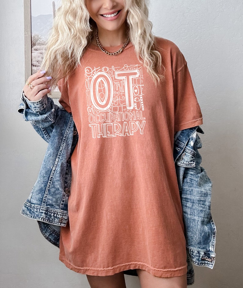 May include: A rust-colored t-shirt with a white graphic design that says "OT Occupational Therapy". The graphic is made up of the letters "OT" surrounded by smaller versions of the letters "OT" and other related words.