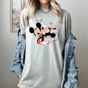 May include: A light green t-shirt featuring a cartoon image of Mickey Mouse and Minnie Mouse kissing. The image is surrounded by small red hearts.