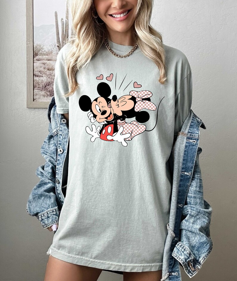 May include: A light green t-shirt featuring a cartoon image of Mickey Mouse and Minnie Mouse kissing. The image is surrounded by small red hearts.