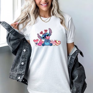 K&ouml;nnte beinhalten: Wei&szlig;es T-Shirt mit einer blauen Comicfigur, die einen Lolli h&auml;lt und von rosa Herzen und S&uuml;&szlig;igkeiten umgeben ist. Die Figur tr&auml;gt ein rotes Stirnband mit den Buchstaben "XO" darauf.