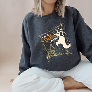May include: Dark gray sweatshirt with the text "I'M A DOG PERSON" and a cartoon illustration of a ghost dog. The design includes a spooky house and decorative elements in gold and orange.