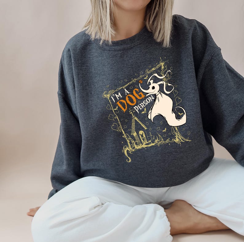 May include: Dark gray sweatshirt with the text "I'M A DOG PERSON" and a cartoon illustration of a ghost dog. The design includes a spooky house and decorative elements in gold and orange.
