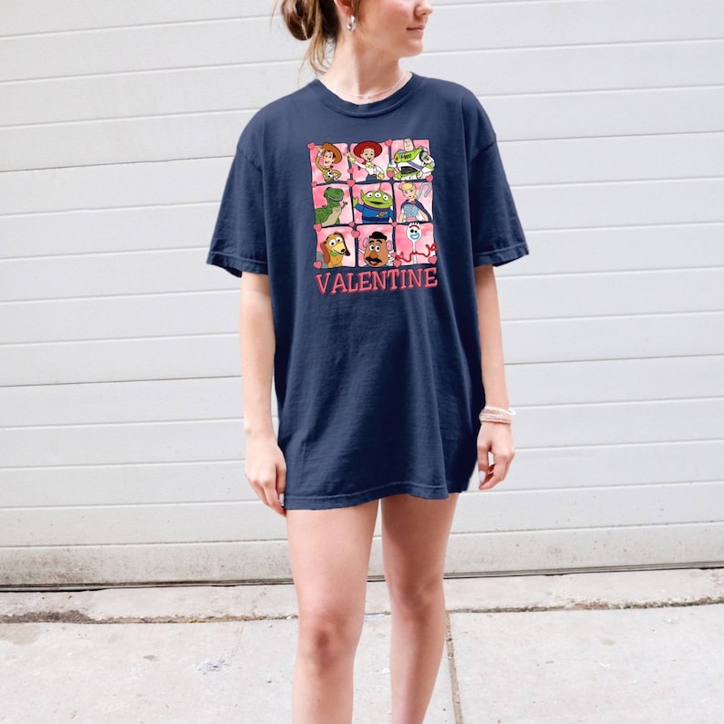 K&ouml;nnte beinhalten: Ein marineblaues T-Shirt mit einem Grafikdruck von Figuren aus der Toy Story-Reihe. Die Figuren sind in einem Raster mit Herzen um sie herum angeordnet. Der Text "VALENTINE" ist unter dem Raster gedruckt.