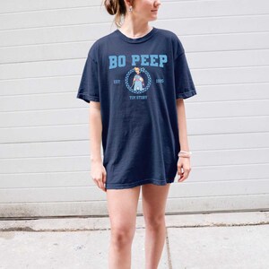 Puede incluir: Una camiseta azul marino con un gr&aacute;fico de Bo Peep de Toy Story. El gr&aacute;fico es un c&iacute;rculo con un patr&oacute;n a cuadros y el texto "Bo Peep", "Est 1995", y "Toy Story".