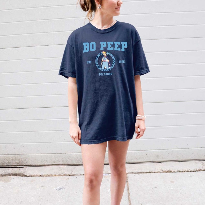 Puede incluir: Una camiseta azul marino con un gr&aacute;fico de Bo Peep de Toy Story. El gr&aacute;fico es un c&iacute;rculo con un patr&oacute;n a cuadros y el texto "Bo Peep", "Est 1995", y "Toy Story".