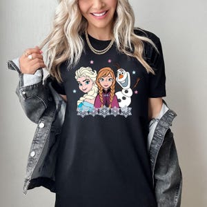 May include: Black t-shirt featuring Elsa, Anna, and Olaf from Frozen. The graphic shows the characters with snowflakes and stars. Elsa has blonde hair, Anna has red hair, and Olaf is a snowman. The shirt is a crew neck.