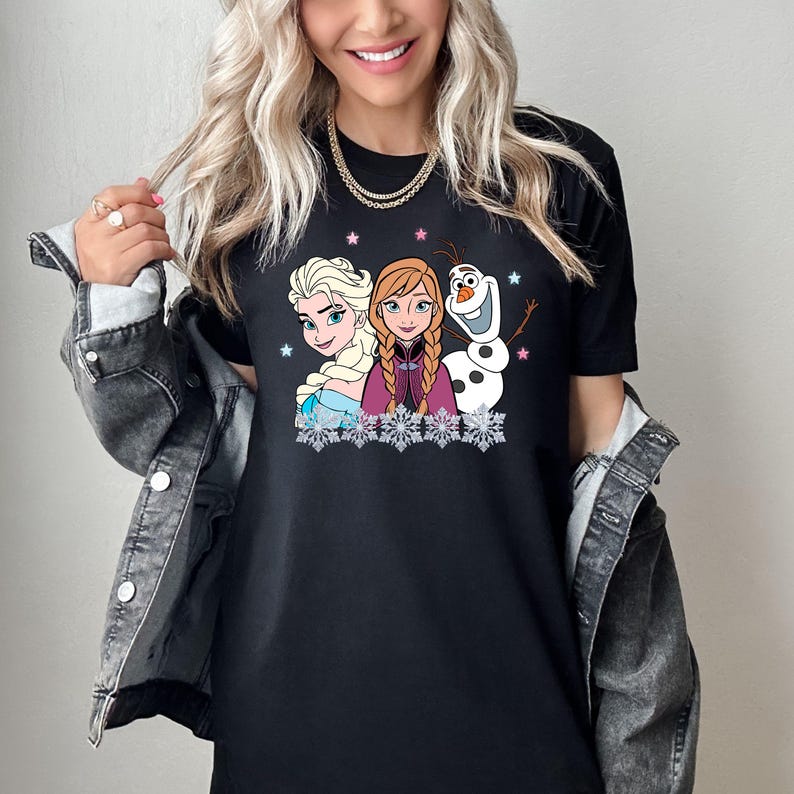 May include: Black t-shirt featuring Elsa, Anna, and Olaf from Frozen. The graphic shows the characters with snowflakes and stars. Elsa has blonde hair, Anna has red hair, and Olaf is a snowman. The shirt is a crew neck.