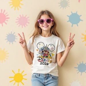 May include: A cream-colored t-shirt featuring a colorful Disney graphic with Mickey and Minnie Mouse, a castle, and the text "Disney 2026." The shirt is worn by a person wearing pink sunglasses and blue jeans.
