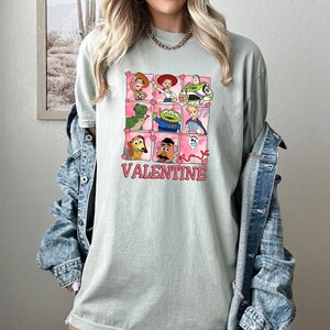 K&ouml;nnte beinhalten: Ein hellgr&uuml;nes T-Shirt mit einem Aufdruck von Toy Story-Charakteren in einem Raster mit Herzen um jeden Charakter. Der Text "VALENTINE" befindet sich unter dem Raster.