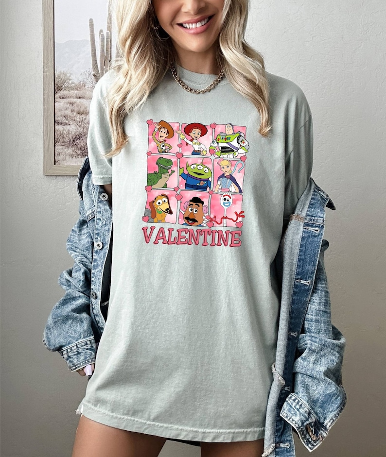 K&ouml;nnte beinhalten: Ein hellgr&uuml;nes T-Shirt mit einem Aufdruck von Toy Story-Charakteren in einem Raster mit Herzen um jeden Charakter. Der Text "VALENTINE" befindet sich unter dem Raster.