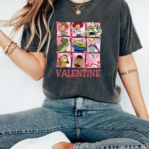 K&ouml;nnte beinhalten: Ein dunkelgraues T-Shirt mit einem Grafikdruck von Figuren aus dem Toy Story-Film-Franchise. Die Figuren sind in einem Raster angeordnet, mit rosa Herzen um jede Figur. Der Text "VALENTINE" ist unter dem Raster gedruckt.