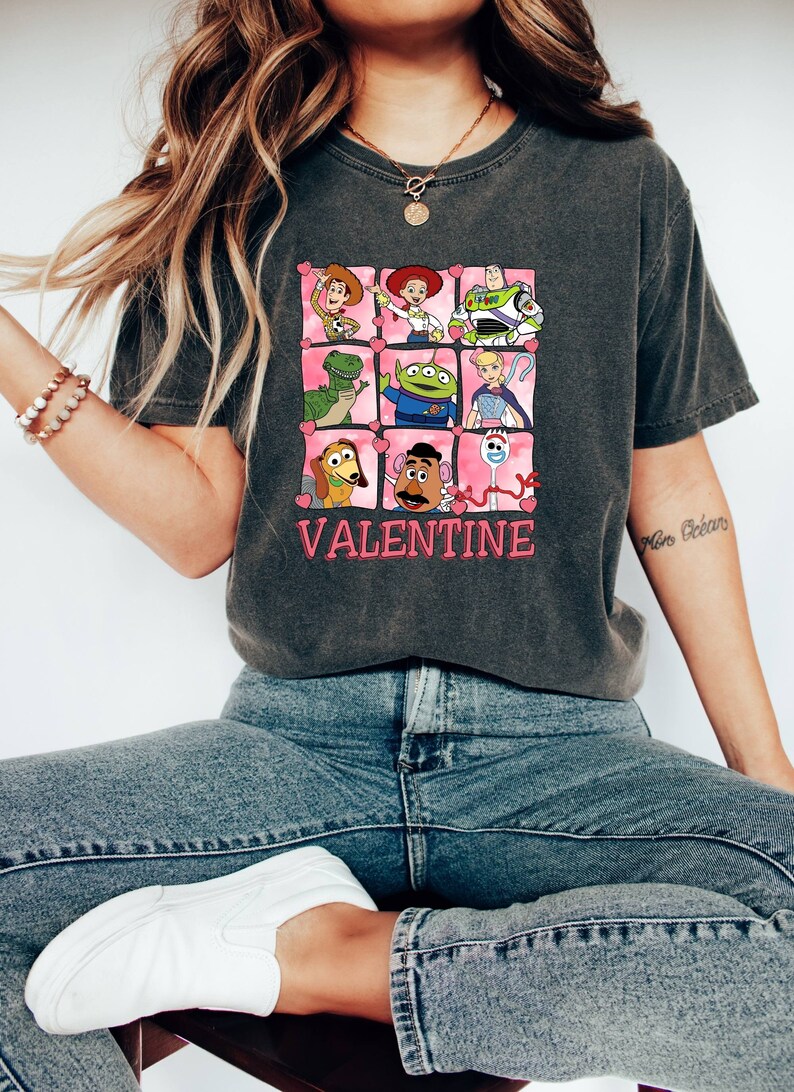 K&ouml;nnte beinhalten: Ein dunkelgraues T-Shirt mit einem Grafikdruck von Figuren aus dem Toy Story-Film-Franchise. Die Figuren sind in einem Raster angeordnet, mit rosa Herzen um jede Figur. Der Text "VALENTINE" ist unter dem Raster gedruckt.