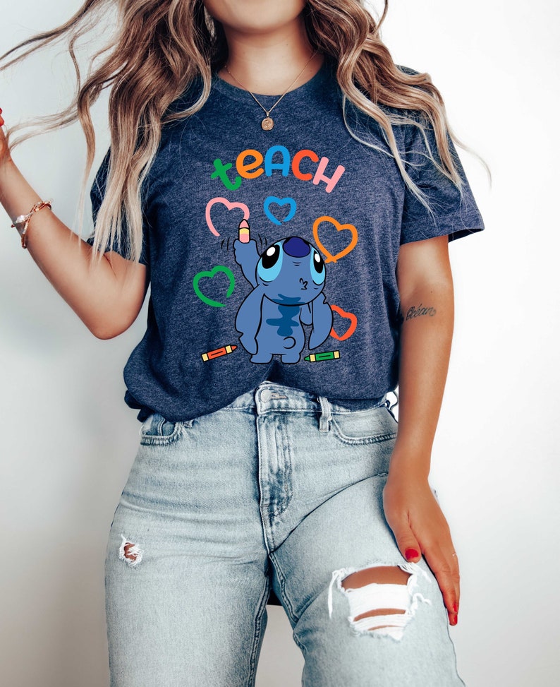 May include: A navy blue t-shirt featuring the word "teach" in rainbow colours and a blue cartoon character holding a pencil. The character is surrounded by colourful hearts and crayons.