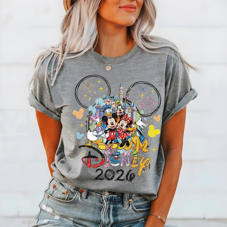 May include: Gray t-shirt featuring a colorful Disney design. The design includes Mickey and Minnie Mouse, Donald Duck, and other characters in front of a castle. The text "Disney 2026" is also visible.