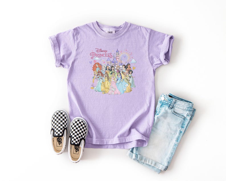 May include: A lilac-colored t-shirt featuring a graphic of Disney Princesses and the text "Disney Princess". The shirt is paired with light blue denim shorts and black and white checkered slip-on shoes.