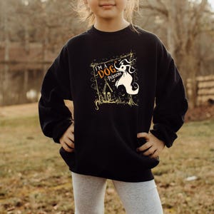 May include: Black sweatshirt with a graphic that reads "I'M A DOG PERSON" with a cartoon dog and haunted house design. The sweatshirt is worn by a person standing outside in front of a lake and trees.