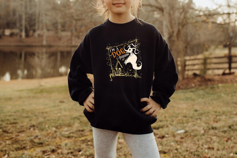 May include: Black sweatshirt with a graphic that reads "I'M A DOG PERSON" with a cartoon dog and haunted house design. The sweatshirt is worn by a person standing outside in front of a lake and trees.