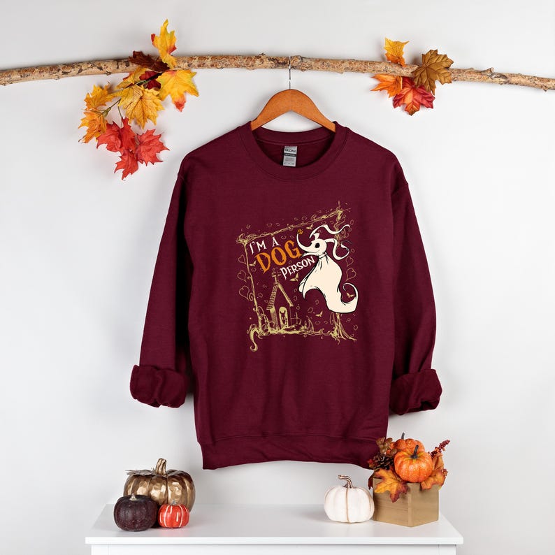 May include: Burgundy sweatshirt with a graphic featuring a ghost dog and the text "I'm a Dog Person." The design is in gold and white. The sweatshirt is hanging on a wooden hanger.
