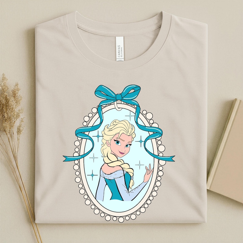 Puede incluir: Camiseta beige con una ilustraci&oacute;n de dibujos animados de una mujer rubia con un vestido azul dentro de un marco ovalado. El marco est&aacute; adornado con una cinta azul y un borde perlado. La camiseta tiene una etiqueta que dice "CANVAS".