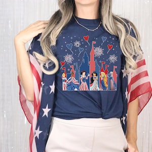 K&ouml;nnte beinhalten: Marineblaues T-Shirt mit einer Schloss-Silhouette, Feuerwerk und herzf&ouml;rmigen Ballons. Das Shirt zeigt Zeichentrickprinzessinnen in patriotischer Kleidung. Ein rot-wei&szlig;-blauer Schal ist &uuml;ber die Schultern drapiert.