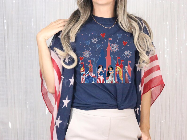K&ouml;nnte beinhalten: Marineblaues T-Shirt mit einer Schloss-Silhouette, Feuerwerk und herzf&ouml;rmigen Ballons. Das Shirt zeigt Zeichentrickprinzessinnen in patriotischer Kleidung. Ein rot-wei&szlig;-blauer Schal ist &uuml;ber die Schultern drapiert.