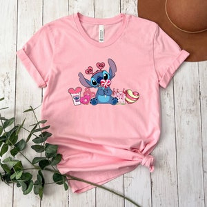 K&ouml;nnte beinhalten: Rosa T-Shirt mit kurzen &Auml;rmeln und einem blauen Stitch-Charakter, der einen Lolli h&auml;lt und von Herzen und S&uuml;&szlig;igkeiten umgeben ist. Das Shirt hat einen Rundhalsausschnitt und ist aus weichem, bequemem Stoff.