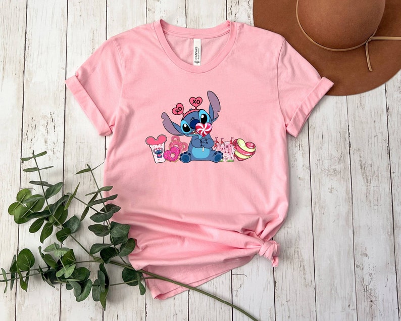 K&ouml;nnte beinhalten: Rosa T-Shirt mit kurzen &Auml;rmeln und einem blauen Stitch-Charakter, der einen Lolli h&auml;lt und von Herzen und S&uuml;&szlig;igkeiten umgeben ist. Das Shirt hat einen Rundhalsausschnitt und ist aus weichem, bequemem Stoff.