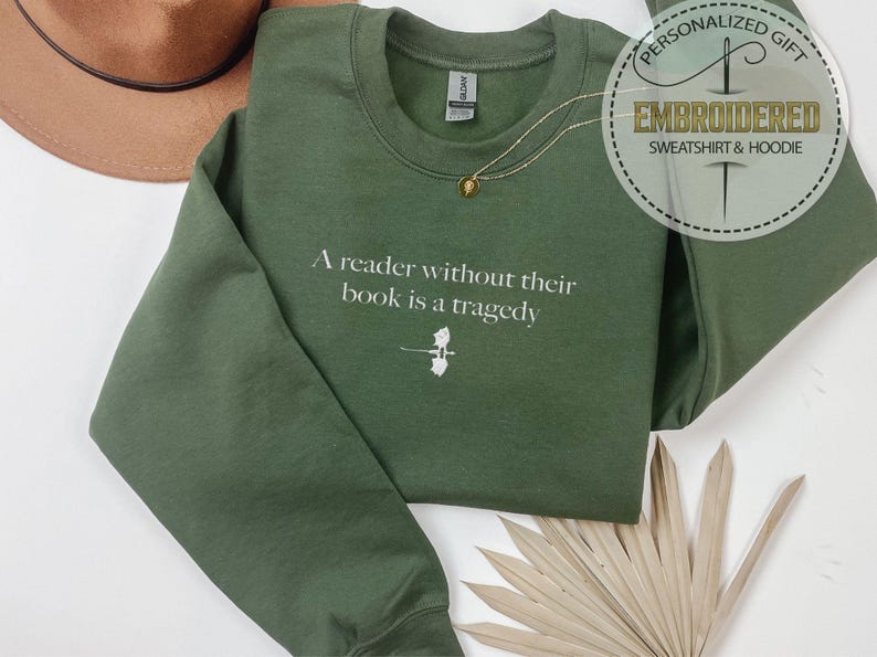 May include: An olive green sweatshirt with the quote "A reader without their book is a tragedy" in white. A small graphic of a book and feather is below the quote. A gold necklace and a brown hat are also visible.