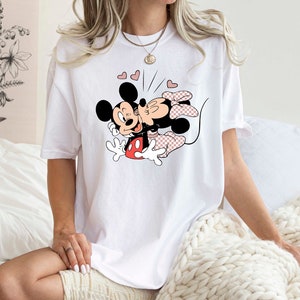 May include: White t-shirt featuring a graphic print of Mickey Mouse and Minnie Mouse kissing. The graphic is in black, red, and pink.