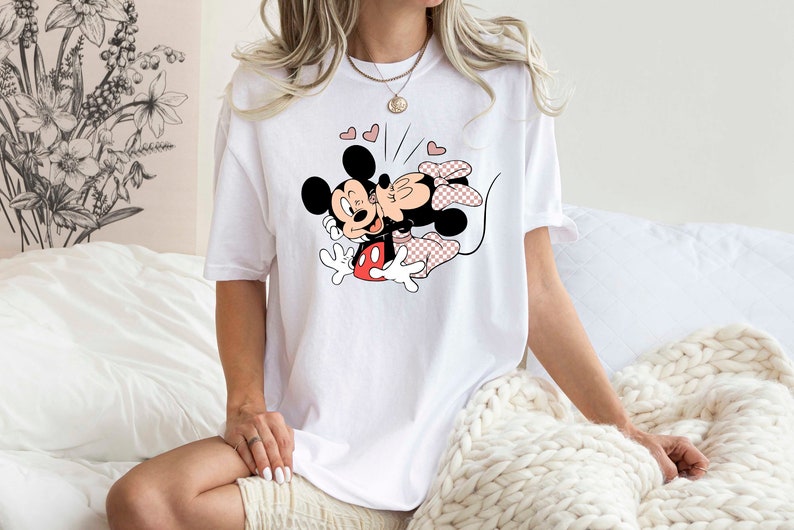May include: White t-shirt featuring a graphic print of Mickey Mouse and Minnie Mouse kissing. The graphic is in black, red, and pink.