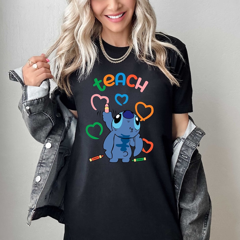 May include: Black t-shirt featuring a blue cartoon character holding a crayon and drawing hearts. The text "teach" is above the character.