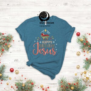 May include: A teal blue t-shirt with a Christmas nativity scene and the text "Happy Birthday Jesus" in gold and pink lettering.