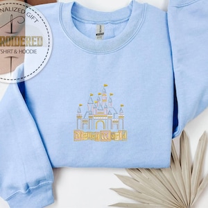 May include: Light blue sweatshirt with an embroidered castle design and the words "Disney World" in gold. The sweatshirt has a crew neck and long sleeves. The image also includes a logo that says "Personalized Gift EMBROIDERED SWEATSHIRT & HOODIE".