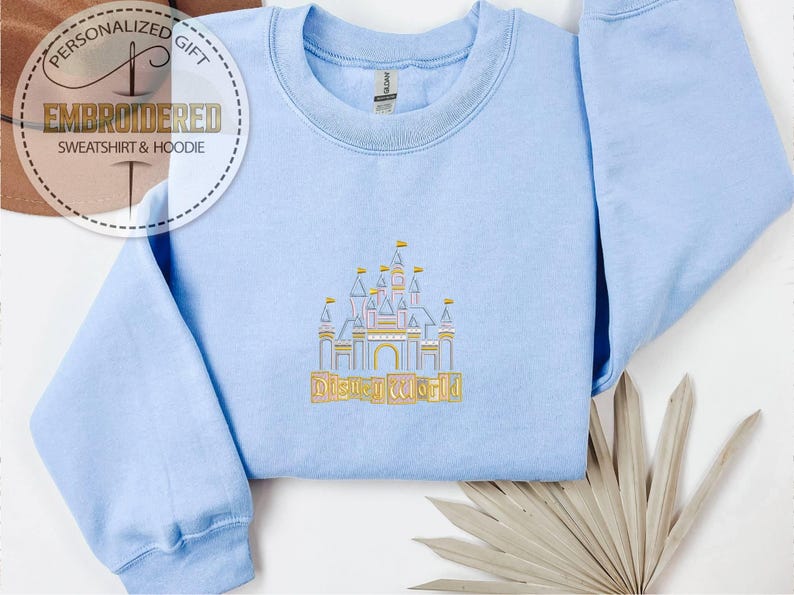 May include: Light blue sweatshirt with an embroidered castle design and the words "Disney World" in gold. The sweatshirt has a crew neck and long sleeves. The image also includes a logo that says "Personalized Gift EMBROIDERED SWEATSHIRT & HOODIE".