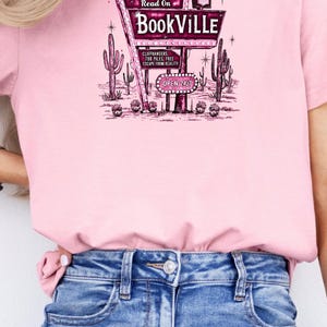 May include: A pink t-shirt with a graphic of a vintage neon sign that reads "Welcome & Read On BookVILLE" with cacti and the text "Cliffhangers, TBR Piles, Free Escape from Reality, Open 24/7".
