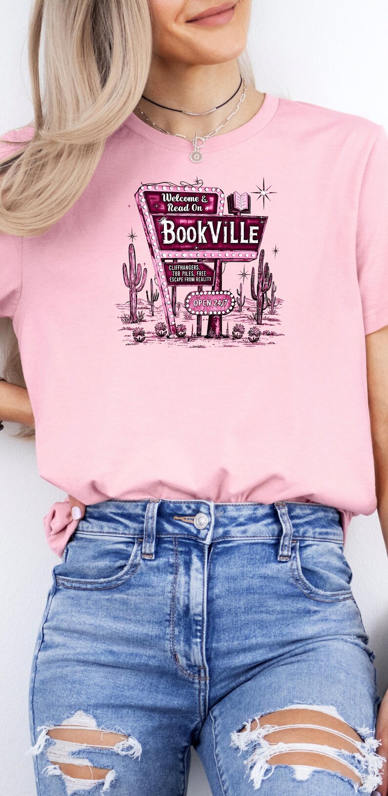 May include: A pink t-shirt with a graphic of a vintage neon sign that reads "Welcome & Read On BookVILLE" with cacti and the text "Cliffhangers, TBR Piles, Free Escape from Reality, Open 24/7".