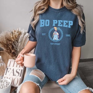 Puede incluir: Una camiseta azul marino con un gr&aacute;fico de Bo Peep de Toy Story. El texto en la camiseta dice "Bo Peep Est. 1995 Toy Story".