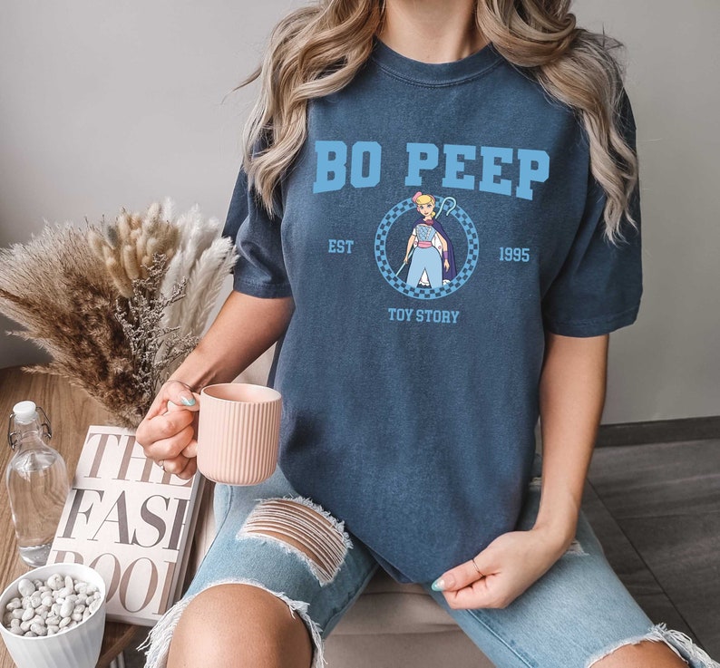 Puede incluir: Una camiseta azul marino con un gr&aacute;fico de Bo Peep de Toy Story. El texto en la camiseta dice "Bo Peep Est. 1995 Toy Story".
