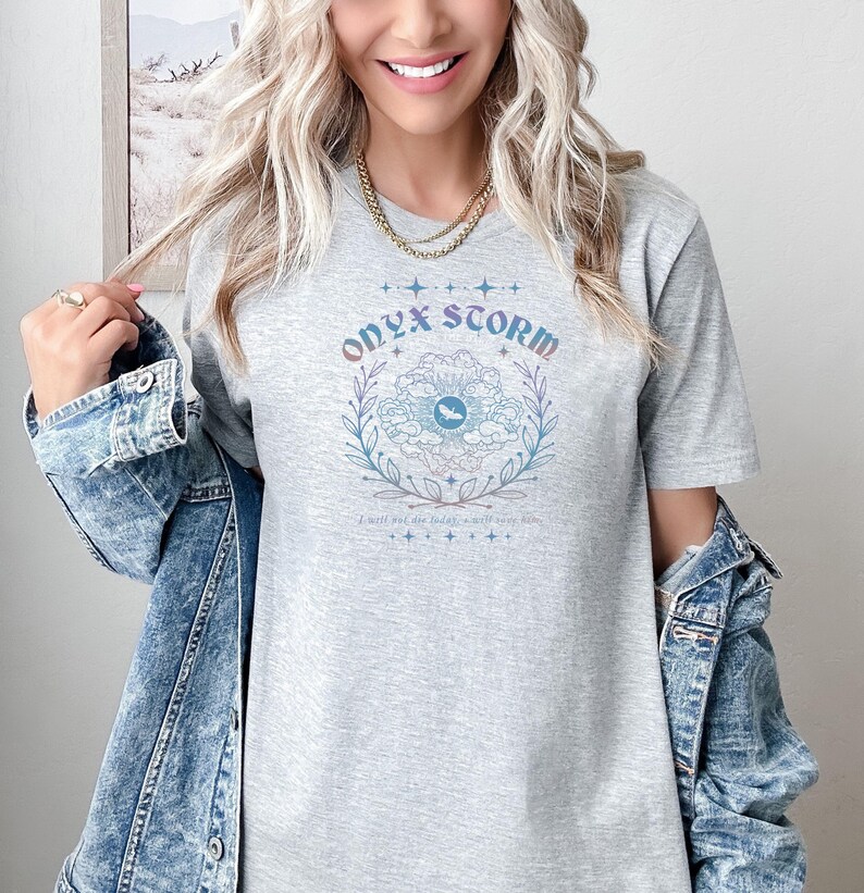 Peut inclure: Un t-shirt gris avec un motif graphique d'un &oelig;il stylis&eacute; avec des nuages et des feuilles. Le texte "Onyx Storm" est au-dessus de l'&oelig;il et le texte "I will put out the fire in my soul" est en dessous de l'&oelig;il.