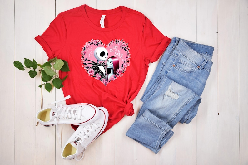 May include: A red t-shirt featuring a graphic of Jack and Sally from The Nightmare Before Christmas inside a pink heart with the text "Love You to Death". The shirt is paired with light blue jeans and white sneakers.