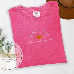 May include: A hot pink Comfort Colors t-shirt with the phrase "Pumpkin Spice and everything nice" embroidered in white and orange. The shirt has a crew neck and short sleeves. A brown hat is visible in the background.