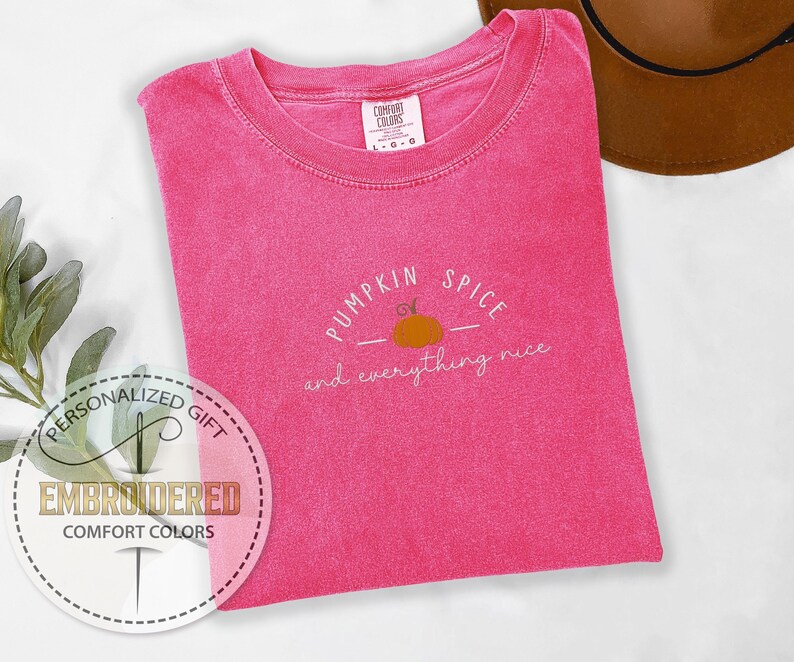May include: A hot pink Comfort Colors t-shirt with the phrase "Pumpkin Spice and everything nice" embroidered in white and orange. The shirt has a crew neck and short sleeves. A brown hat is visible in the background.
