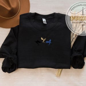 May include: Black sweatshirt with embroidered dragon silhouettes in gold, blue, and black. The sweatshirt has a crew neck and long sleeves. The image also includes a brown hat, wheat stalks, and a circular logo that says "PERSONALIZED GIFT EMBROIDERED SWEATSHIRT & HOODIE."