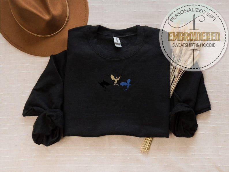 May include: Black sweatshirt with embroidered dragon silhouettes in gold, blue, and black. The sweatshirt has a crew neck and long sleeves. The image also includes a brown hat, wheat stalks, and a circular logo that says "PERSONALIZED GIFT EMBROIDERED SWEATSHIRT & HOODIE."